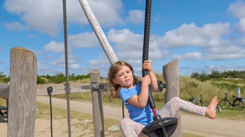 A child happily jumps on a zip wire in a wide, sunny playground setting with a cloudy, blue sky