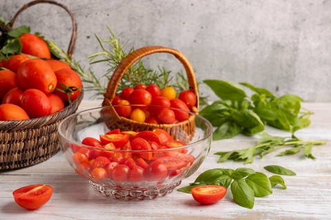 Sliced tomatoes in a glass bowl, surrounded by fresh tomatoes in baskets, basil and rosemary
