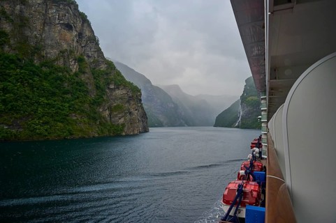 A cruise ship, my ship 6, sails through a misty fjord surrounded by green mountains and waterfalls,