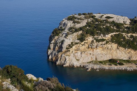 Steep rocky coast with calm, blue water and green vegetation under a clear sky, Anthony Quinn Bay,