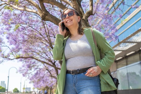 A young woman in casual attire talks on her smartphone while sightseeing by jacaranda trees and a