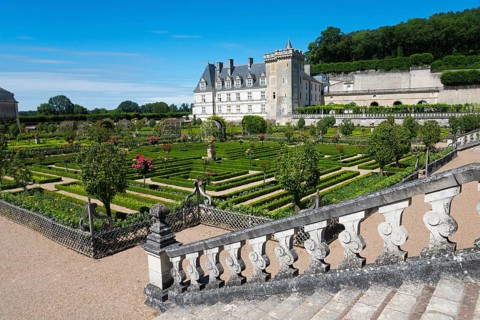 A castle with a magnificent, symmetrical garden full of flowers and shrubs under a clear blue sky,