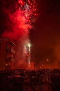 Fireworks explode at night over a residential building with smoke billowing in the background,