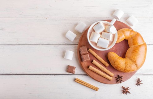 A cup of hot chocolate with marshmallow, croissant and spices on white wooden background. top view,