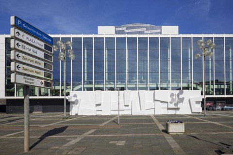 Front of the Musiktheater im Revier in front of a blue sky with lanterns and signs of the parking