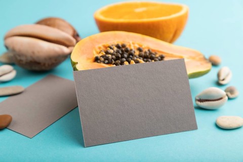 Gray paper business card with ripe cut papaya, orange, seashells on blue pastel pastel background.