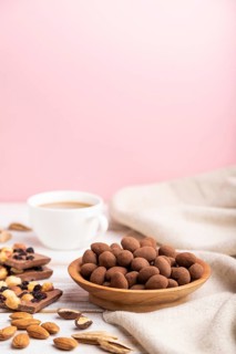 Almond in chocolate dragees in wooden plate and a cup of coffee on white and pink background and