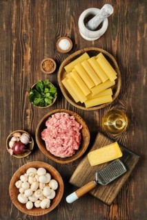 A variety of fresh ingredients is arranged on a wooden surface, featuring ground meat, pasta