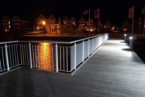 Pier with illuminated lamps, railings and light reflections in the water at night, flags and houses