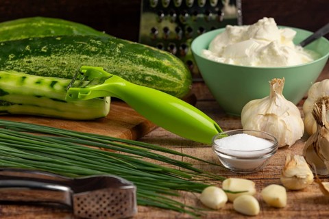 Fresh ingredients such as cucumbers, garlic, chives and sour cream on a wooden table with a peeler