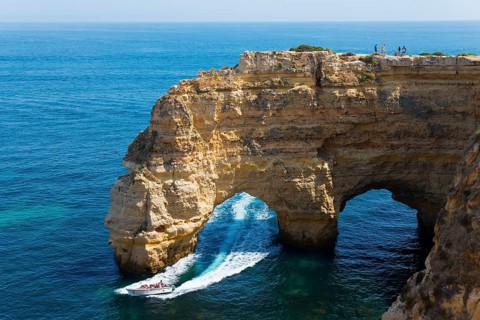 A boat sails under a natural rock arch along an impressive rocky coast, Praia da Marinha, double
