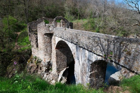 Alte Steinbrücke über einen Fluss, umgeben von bewaldeter Landschaft und Ruinenresten im Frühling,
