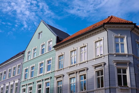 Several old buildings with different facade colours and red roof tiles in front of a blue sky with