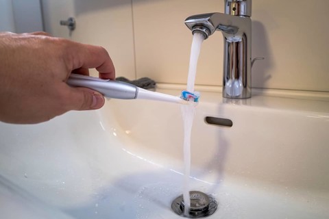 A person holds an electric toothbrush under a running tap in the bathroom, symbolic image of water