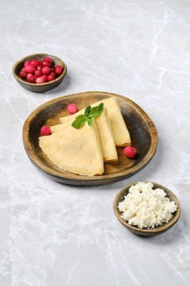 Three golden crepes are elegantly arranged on a wooden plate, accompanied by small bowls of fresh
