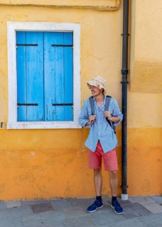 Young man with backpack in front of yellow house wall, yellow house facade with windows with blue
