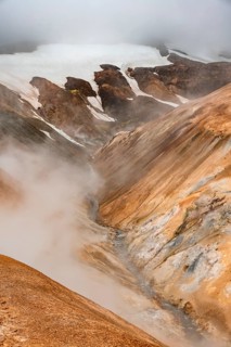 Steaming stream between colourful rhyolite mountains with snowfields, Hveradalir geothermal area,
