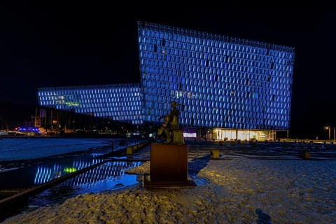 Bronze statue of the man with the cello, behind it the illuminated facade of the concert hall and