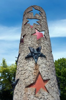 Column with punched-out stars and metal reliefs under a blue sky, National Column in front of the