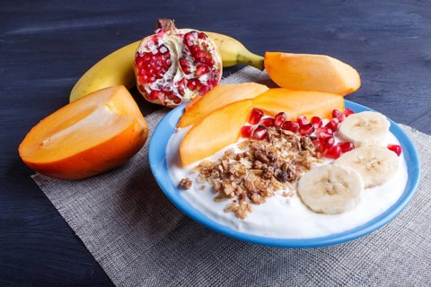 A blue plate with greek yogurt, granola, persimmon, banana, pomegranate black wooden background.