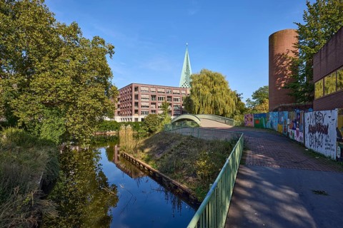 River Bocholter Aa with footpath, trees and pedestrian bridge under a cloudless blue sky in