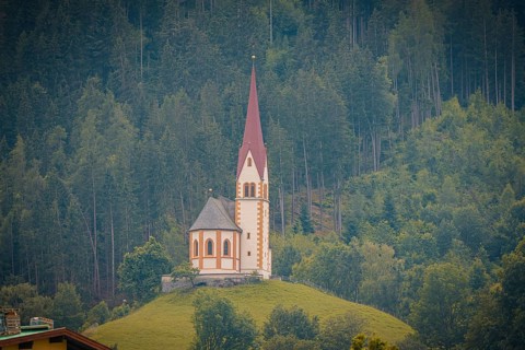 A church with a high tower stands on a green hill in front of a dense forest, Mayrhofen, Zillertal,