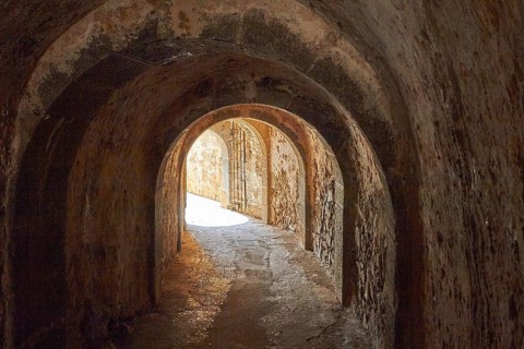 View through a dark tunnel with arches and stone walls leading to a light-coloured exterior