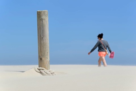 Boundary post on the beach, rope blown over by sand, person crossing the sand drifts, blue sky,