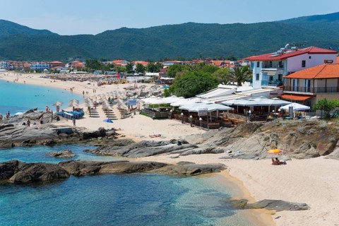 Beach with umbrellas and beach houses, clear blue water and mountains in the background, Sarti,
