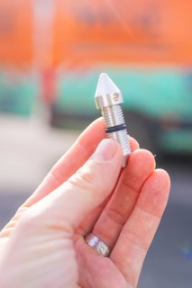 Hand holding a small metal component in front of a blurred background, Wend Glasfasermontage during