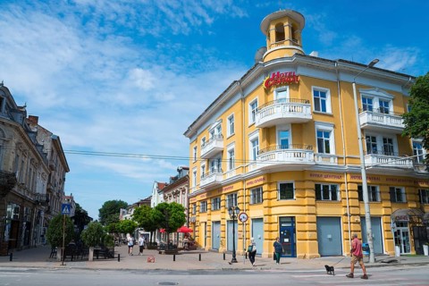 A yellow hotel building under a light blue sky with few clouds and passers-by on the street, hotel