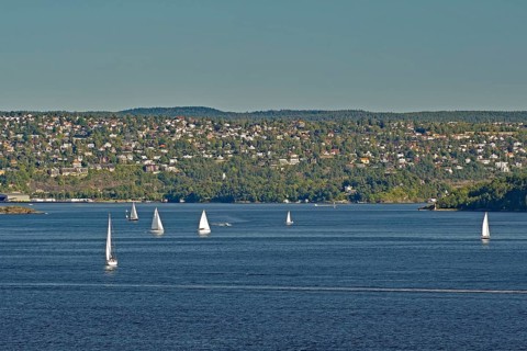 A few sailboats float calmly on the water, in front of a hilly cityscape under a clear sky,