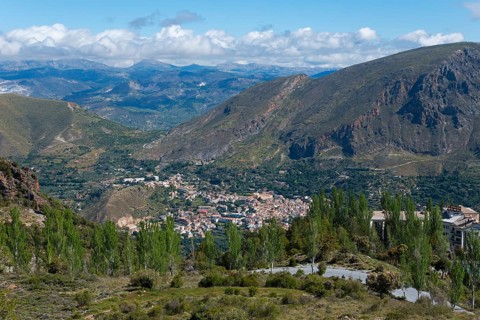 View over a valley with a village surrounded by mountains, lots of greenery and a cloudy sky,