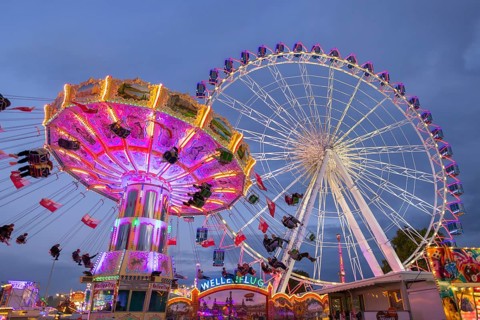 A lively funfair at dusk, illuminated by a large Ferris wheel and a colourful chain carousel,