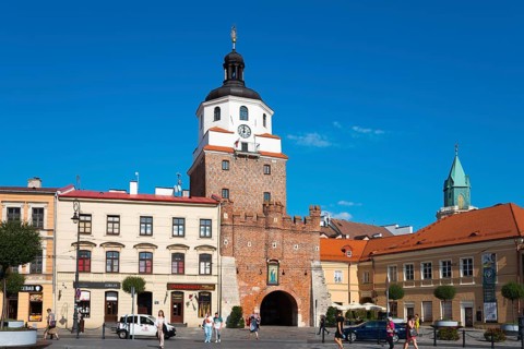 Historic city gate with brick tower and turrets in front of a lively square with passers-by, Krakow