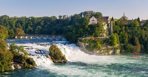 Waterfall flowing next to a historic bridge with lush vegetation and buildings in the background,