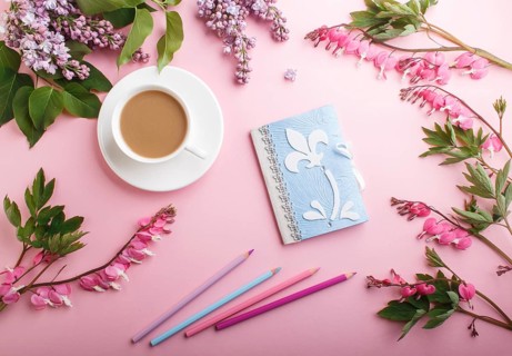 Purple lilac flowers and a cup of coffee with notebook and colored pencils on pastel pink