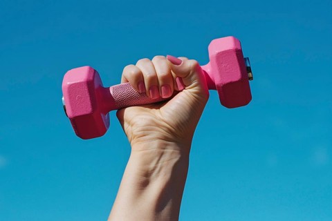 Woman's hand with pink nail polish holding pink dumbbell free weight in front of blue sky.