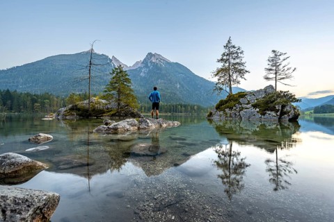 Young man standing on stones in the lake, Hochkalter reflected in Hintersee, at sunset,