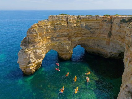 A rocky cliff juts out into the turquoise sea, where several colourful kayaks paddle on the water,