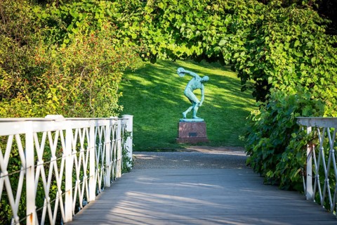 Wooden bridge leading to the sculpture of a discus launcher in a green, sunny park area, Botanical