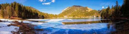 Mountain lake partly covered with snow, surrounded by forests and mountains under a blue sky,