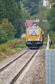 A yellow train runs on rails through a wooded landscape, surrounded by trees and accompanied by