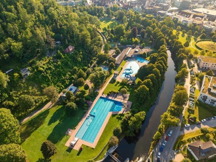 Spacious view of an outdoor swimming pool in the middle of a park, adjacent to a narrow river and