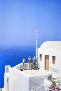 Three male men tourists look out to sea from terrace of whitewashed building at Oia, Santorini,