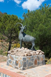 Bronze ibex statue on a brick base surrounded by green trees, Área Recreativa de Sedella, Parque