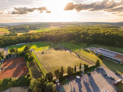 Aerial view of a sports field with football pitch and tennis courts, surrounded by forest and