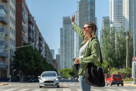 Young woman with sunglasses and a shoulder bag is hailing a ride on a city street, holding a