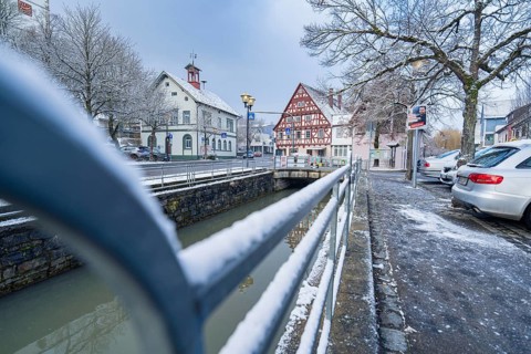 Road with snow cover next to river, view of half-timbered houses and bridge in wintry cityscape,
