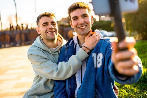 Two men in love are hugging and taking a selfie with a smartphone, enjoying a sunny day outdoors
